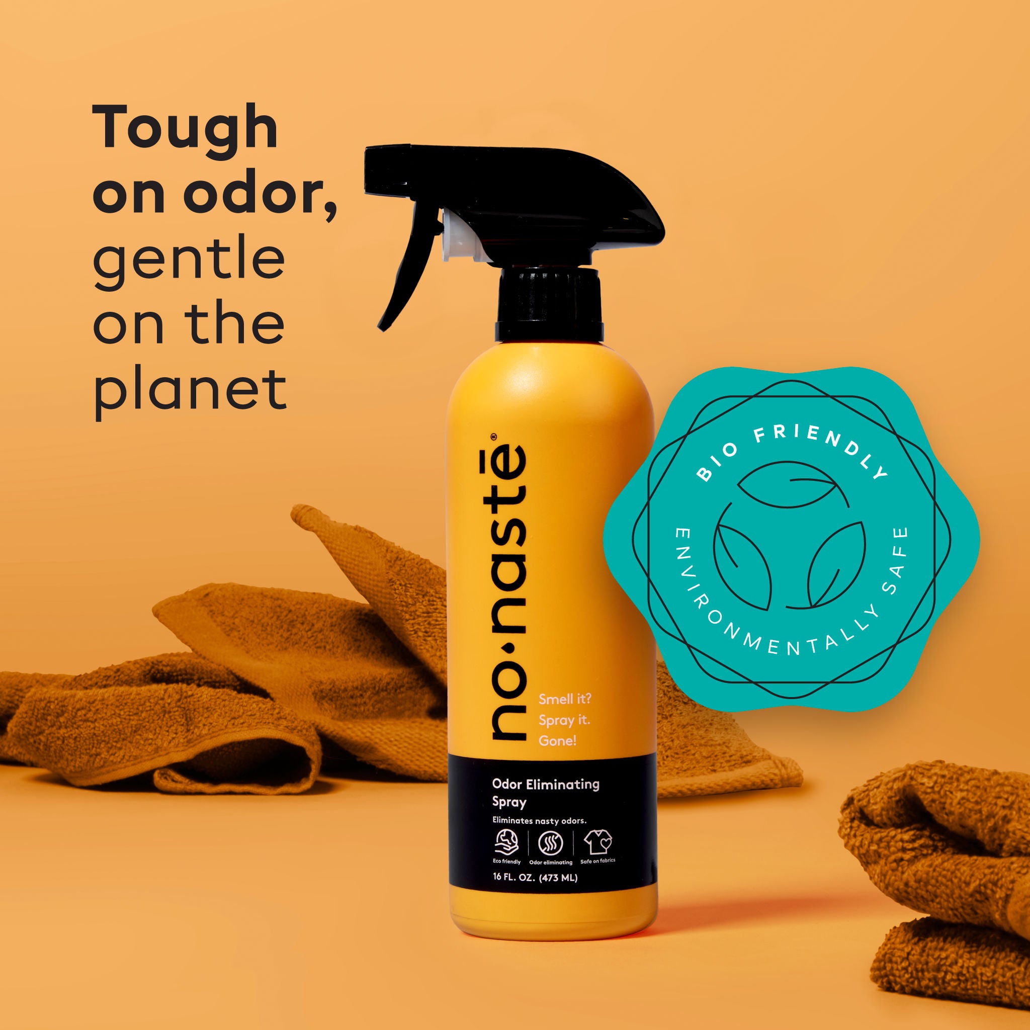 Orange spray bottle with black nozzle labeled 'nonaste' on a beige background with towels and a green eco-friendly logo.